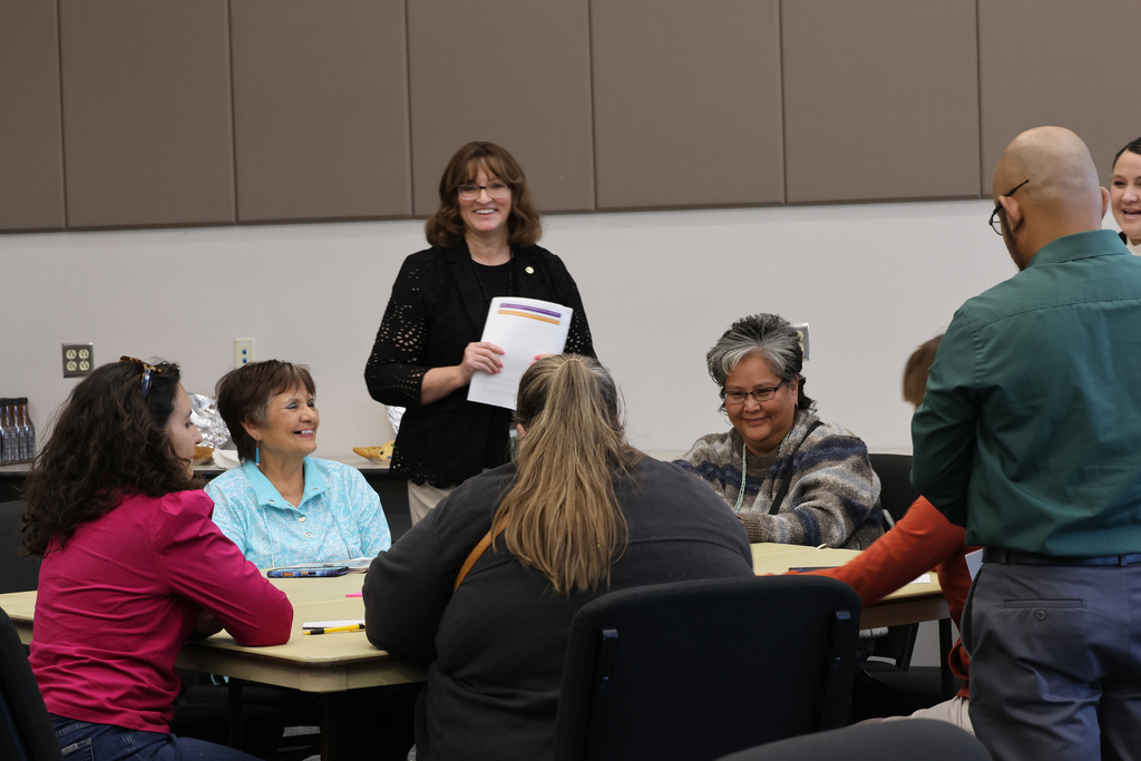 Group of adults sit around a table. A woman in black stands and holds papers.
