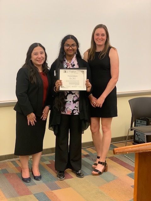 Three women in formal attire stand in a room, one holding a framed certificate. Behind them, a white wall and a wooden podium are visible.
