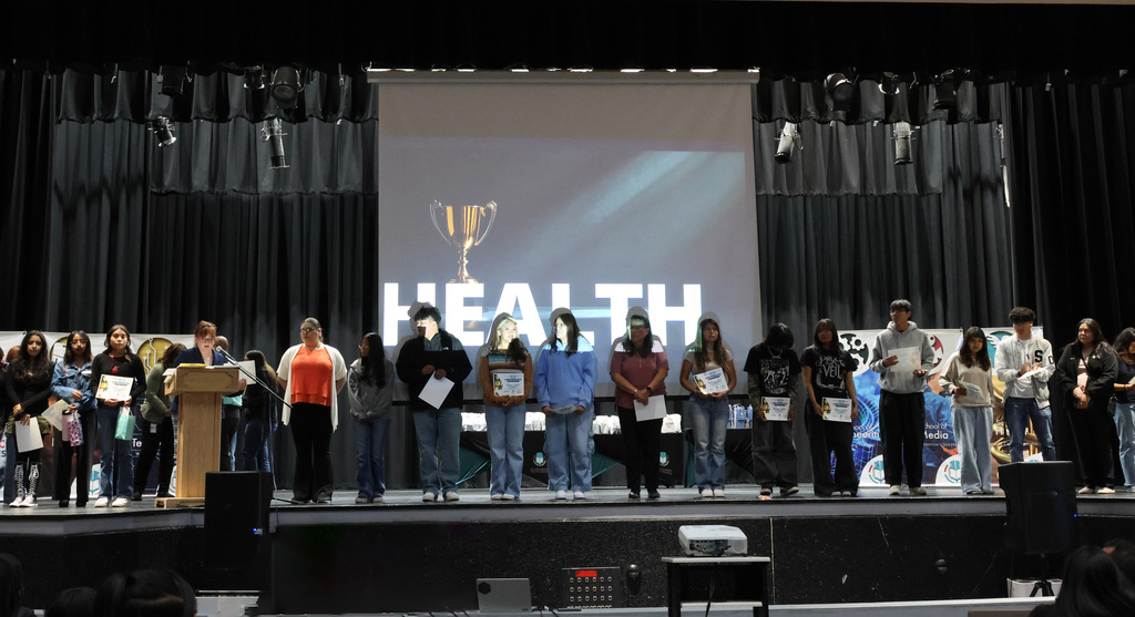 Many people are on a stage holding certificates. A large screen behind them says HEALTH.