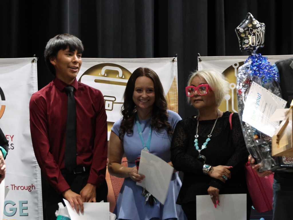 Three people stand together, holding papers. One holds a trophy. Behind them are banners with logos.