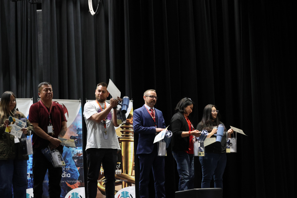 Five people on stage, holding certificates and white papers. Three hold a trophy. Two banners behind them.