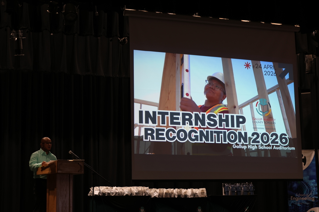 A man stands at a podium in front of a screen displaying a photo of an intern.