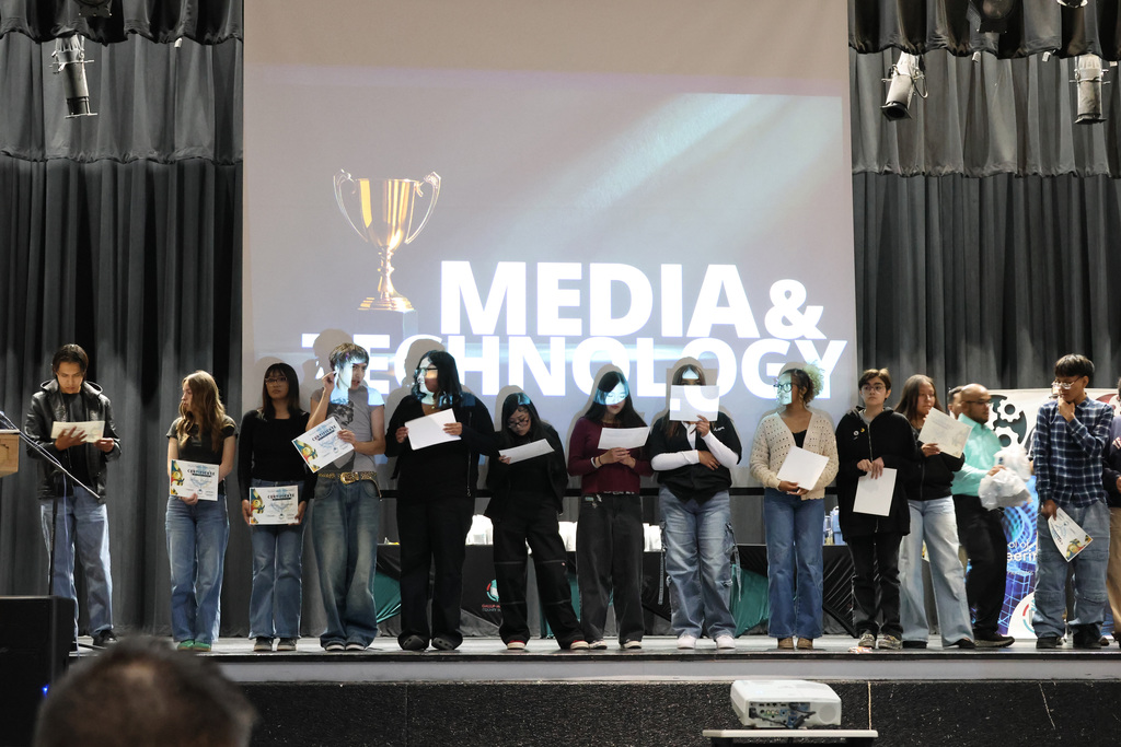 Group of students stand on stage, holding certificates, with a screen displaying "MEDIA & TECHNOLOGY" and a trophy.