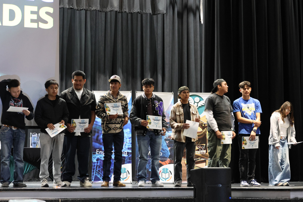 Nine young men on stage holding certificates. A banner behind them reads "GRADES". Two women stand to the right.