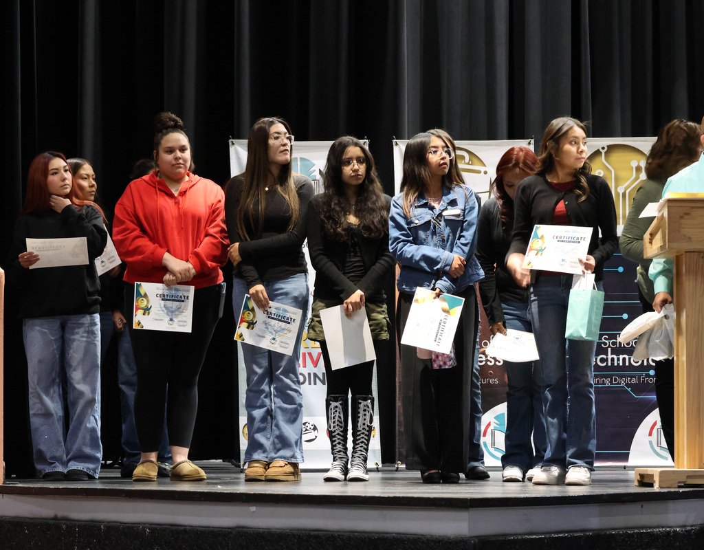 Several women stand on a stage holding papers, likely awards, in front of banners. They wear casual attire.