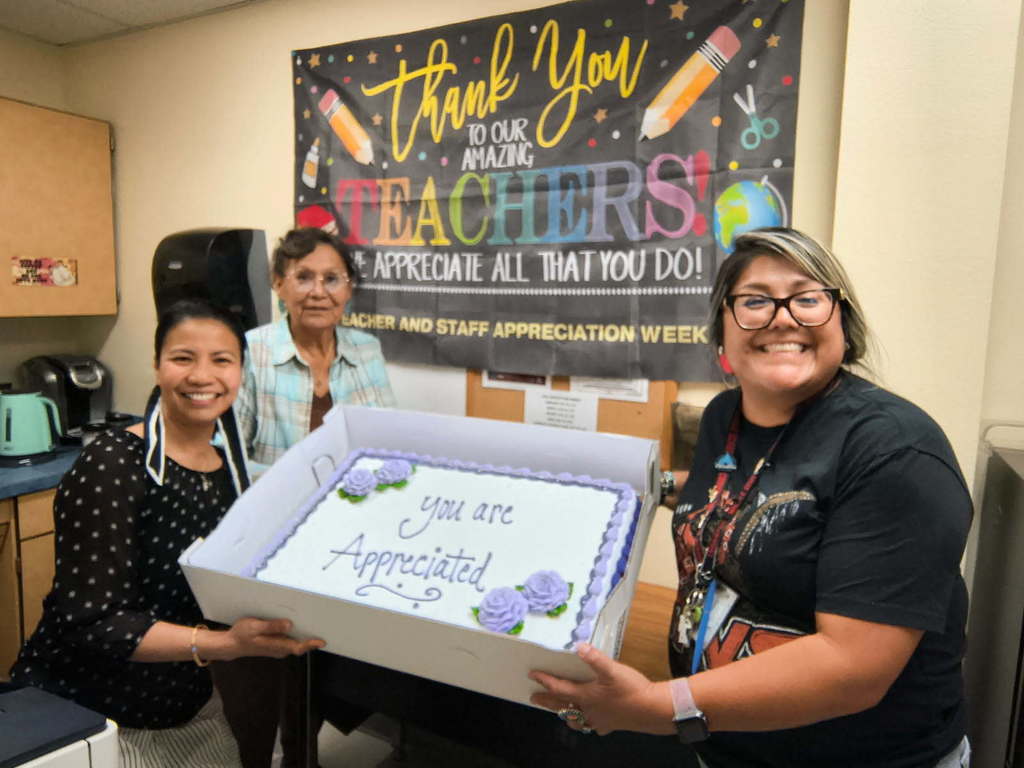 Three people holding a cake in a kitchen. A banner behind them says "Thank You Teachers". One cake says, "You Are Appreciated".