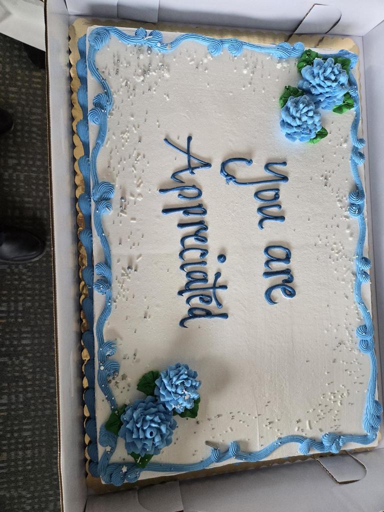A white cake in a box with blue flowers. It reads "Appreciate You. You are." Blue and white icing decorates the cake.