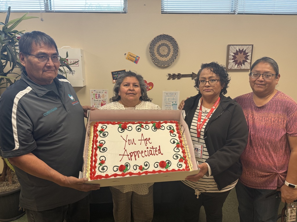 Four people stand together holding a cake with icing that reads, "You Are Appreciated." They are in a room with a potted plant and artwork on the wall.