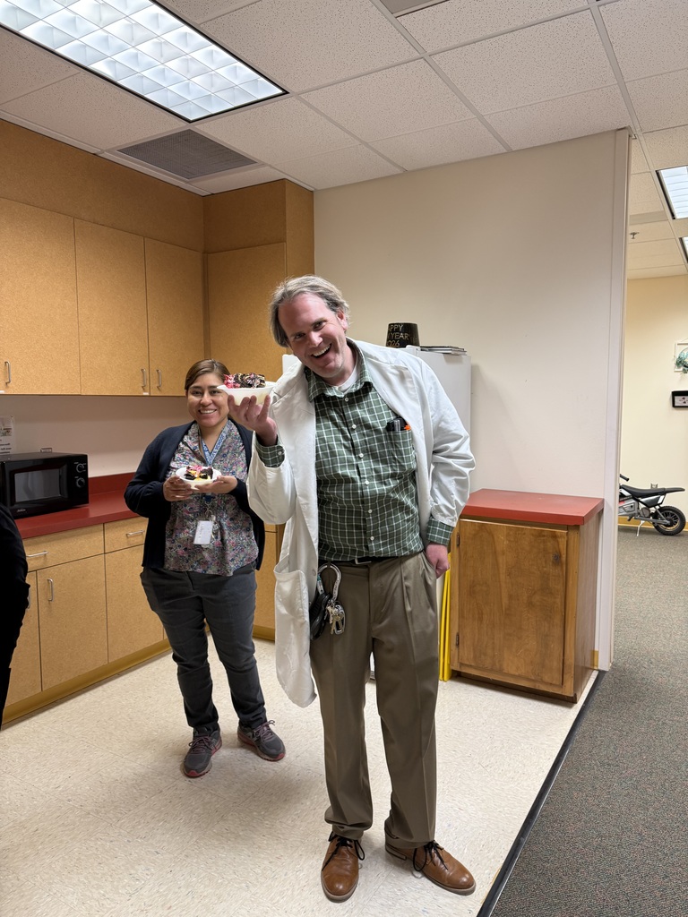 A man and a woman stand in a kitchen area. The man is smiling and holding food. They are in a room with cabinets and a microwave.