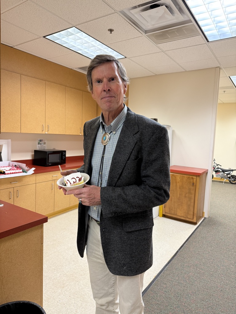 A man in a gray blazer and white pants holds a bowl with food inside a room with wooden cabinets.
