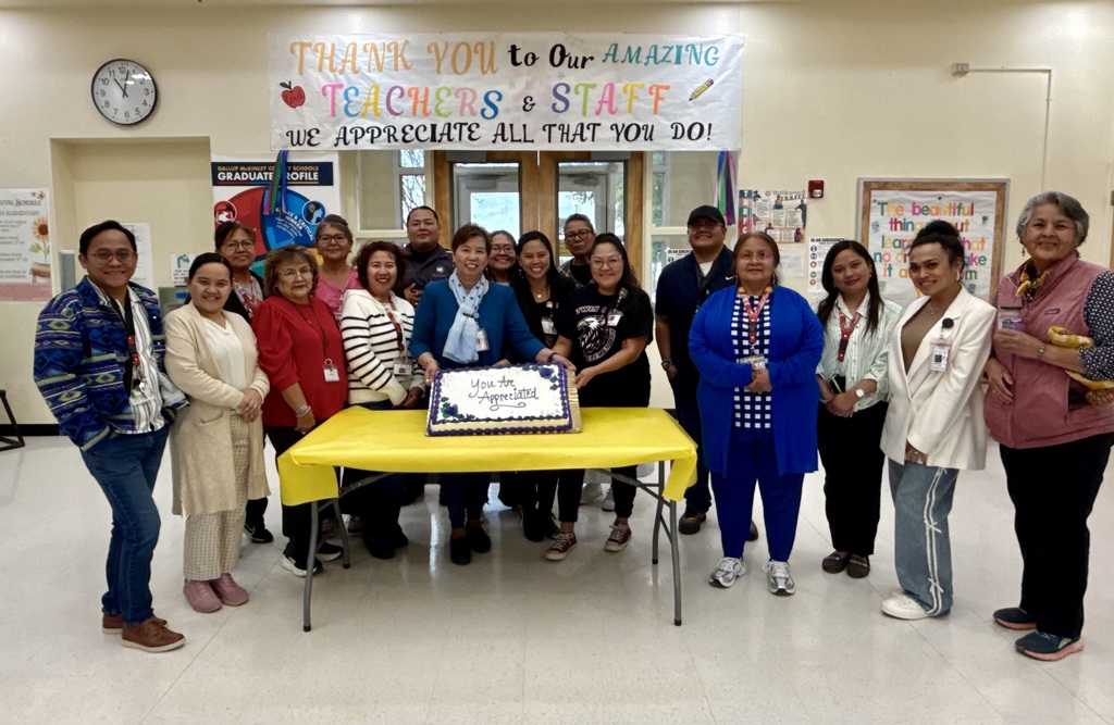 A group of people stand around a table with a cake in a room. A banner reads "Thank you to our amazing teachers and staff."