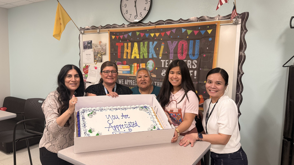 Five women hold a cake box in front of a table with a "Thank You" board. A clock and flags are on the wall.