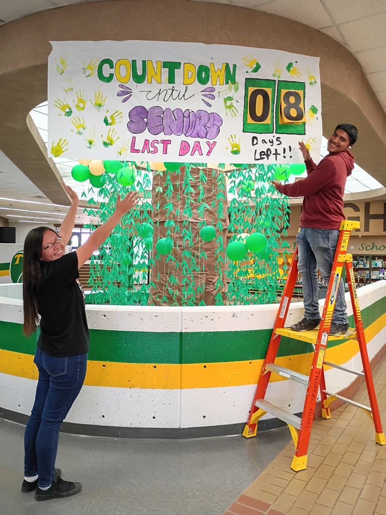 Two people work on a large banner in a school hallway. A woman holds a balloon while a man on a ladder adds more balloons.