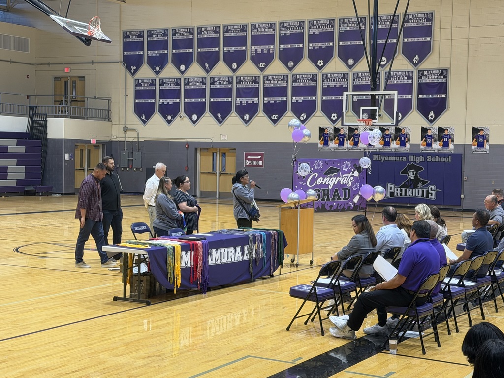 People gathered in a gymnasium with purple banners, standing and seated. A person is speaking at a podium with a microphone.
