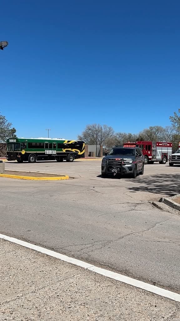 A parking lot with a green and yellow school bus, a black SUV, and two fire trucks under a clear blue sky.