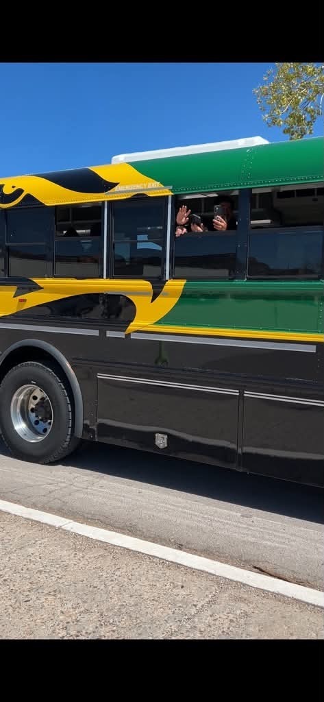 A black and green bus drives on a road. A person inside waves from a window.