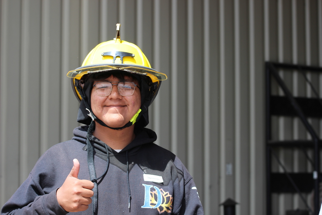 Person in yellow helmet and glasses gives a thumbs-up. Wearing a blue hoodie. Background: gray panels.