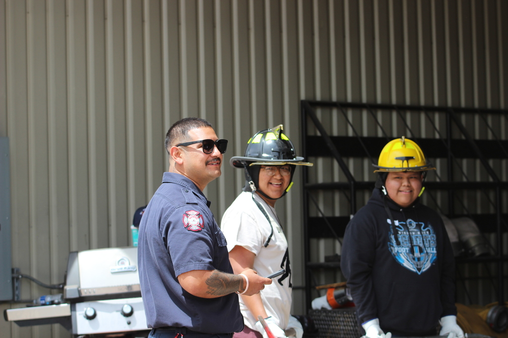 Three people stand in front of a building. One man wears sunglasses and a blue shirt, while the others wear white shirts and helmets.