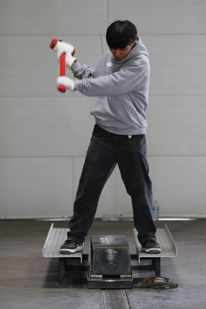A person stands on a metal platform, holding a red hammer in both hands. They wear a gray hoodie, dark pants, and sneakers. A white wall is in the background.