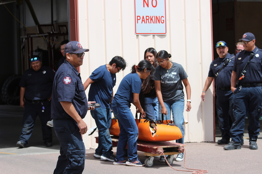 Emergency workers transport equipment on a cart. A red "No Parking" sign hangs above them.