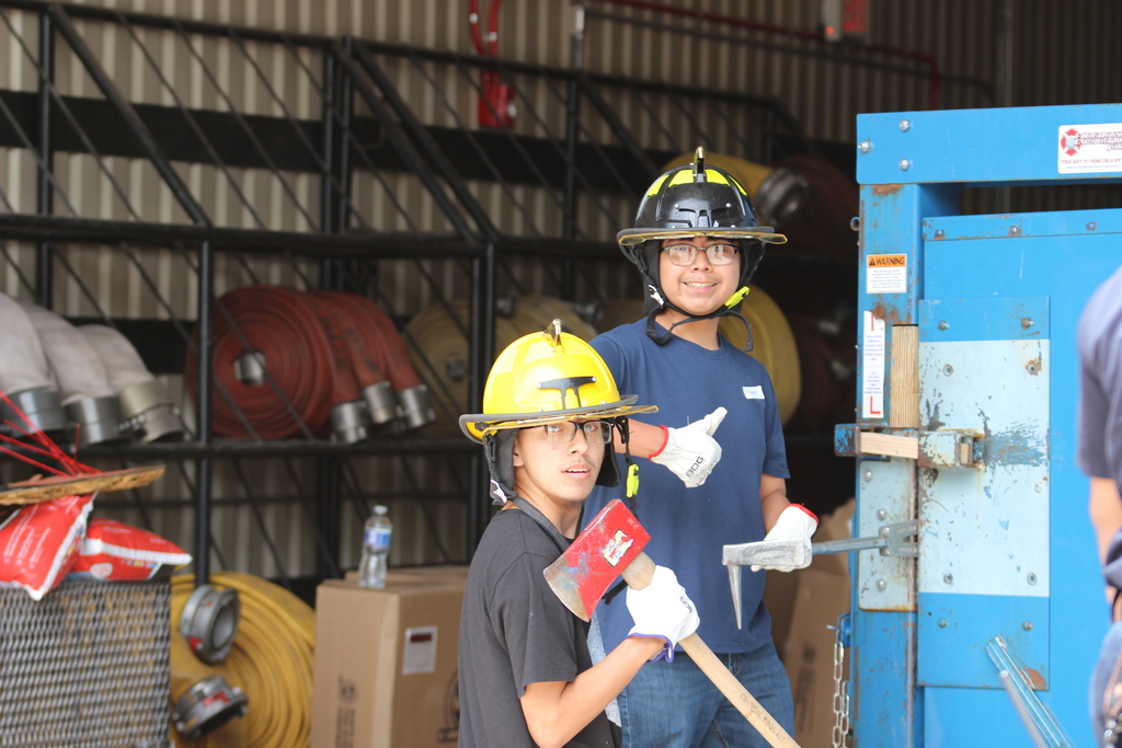 Two people in protective gear stand near a blue door. One holds a sledgehammer, while the other wears a fireman's helmet.