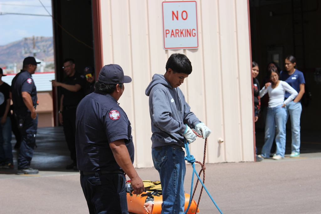 People stand around a building with a "No Parking" sign. Two individuals face each other, one holding a rope.