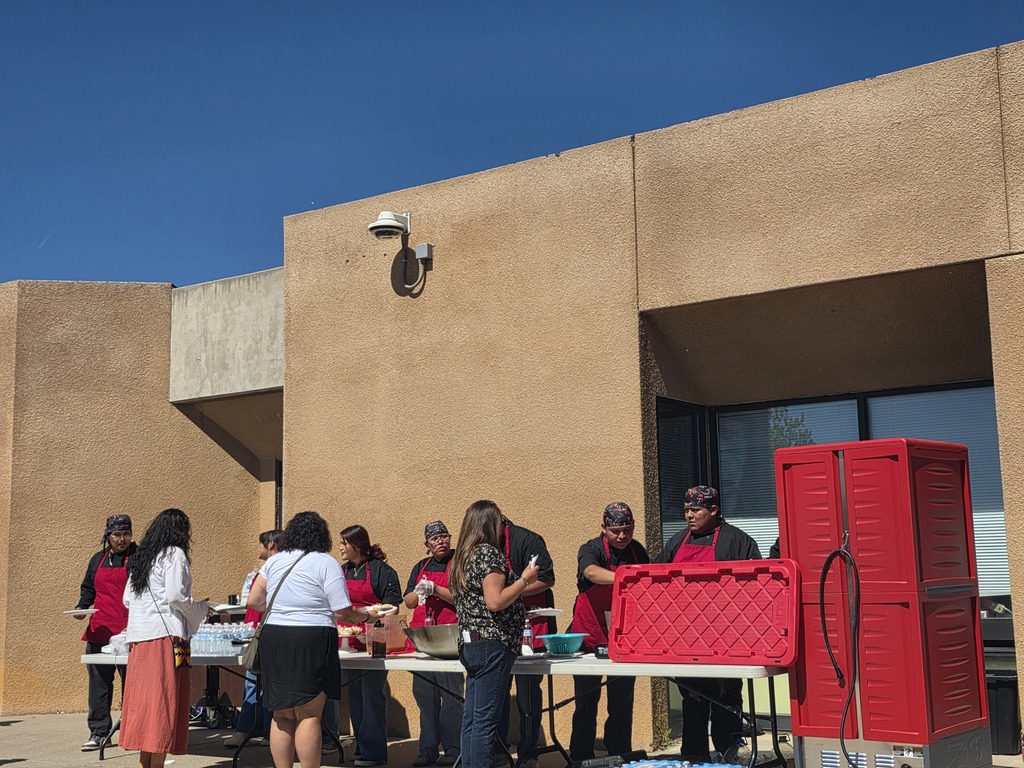 A group of people wearing aprons stands around tables outside a building with a red storage box.