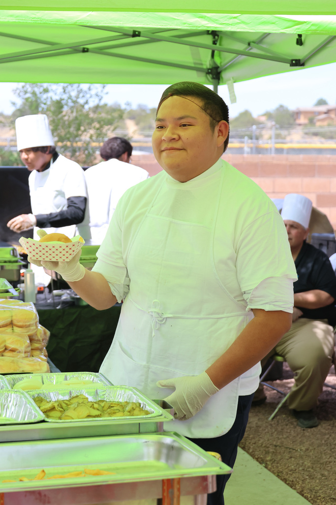 A chef under a green tent, wearing a white uniform and gloves, holding a plate of food.