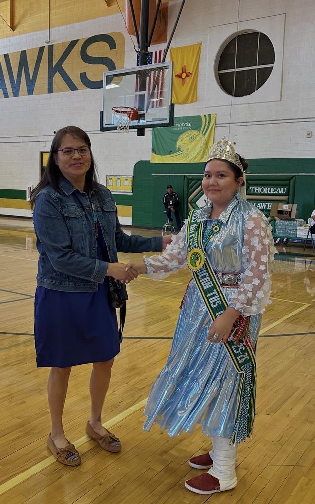 Two women stand in a gymnasium. One wears a blue dress and denim jacket; the other in a blue dress with a sash.