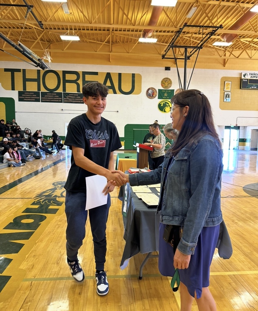 A young man and woman are shaking hands in a gymnasium. He holds a paper, and she wears a denim jacket.