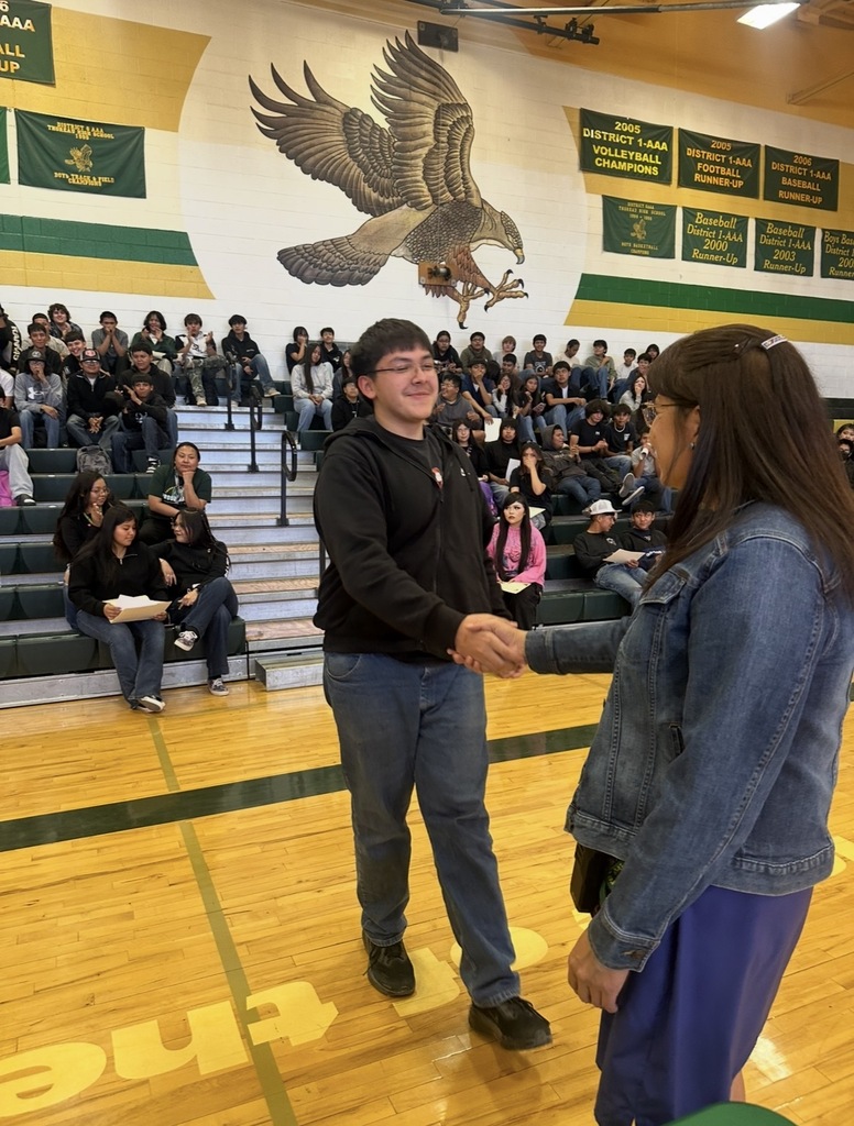 Two people shake hands in a gymnasium. A crowd of people are seated on bleachers. An eagle is on the wall behind them.