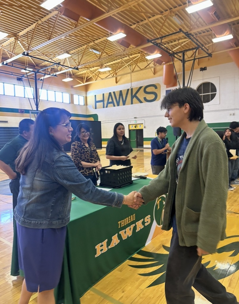 Two people shake hands in front of a table in a gymnasium. A banner reads Hawks.