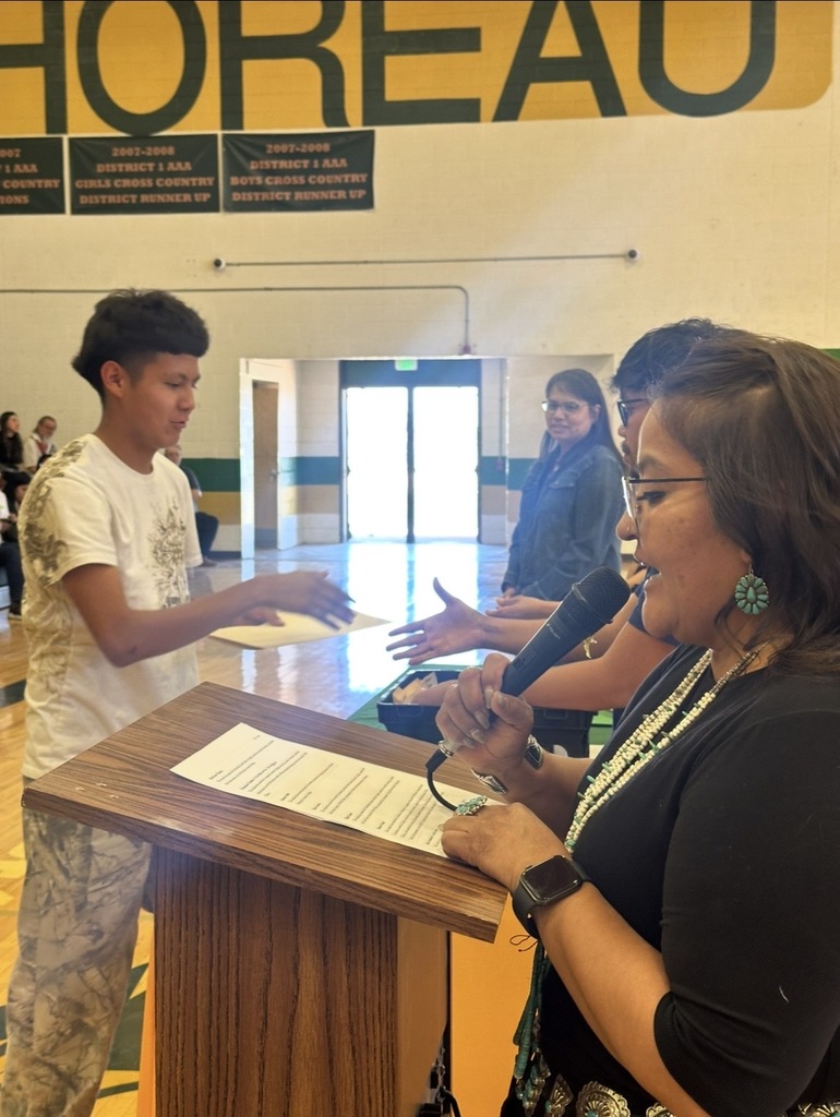 A woman stands at a podium, holding a microphone, and speaking to a boy who is gesturing. Behind them, two other individuals are visible. Banners hang on the wall.