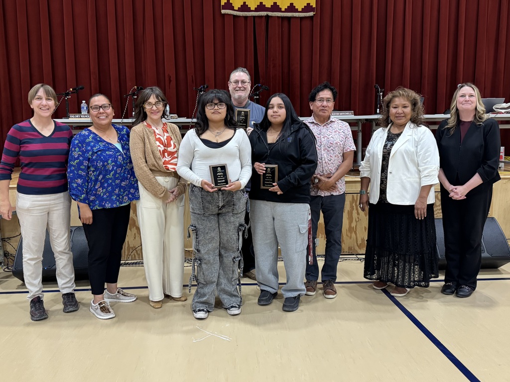 A group of adults stands together in a gymnasium, some holding awards, with a red curtain in the background.