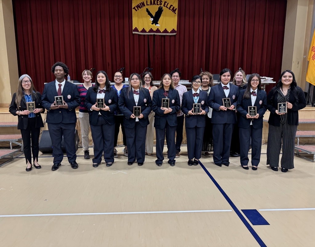 People stand in a row, holding trophies, in a room with red curtains, a banner, and a blue line on the floor.