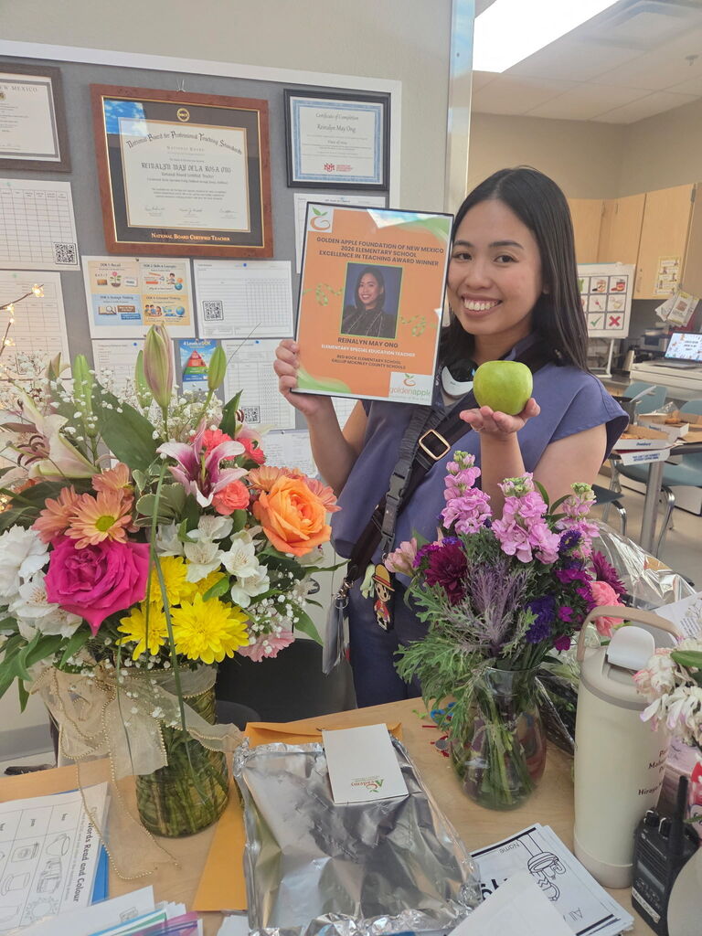 A person holds an orange booklet and an apple over a table with flower bouquets.