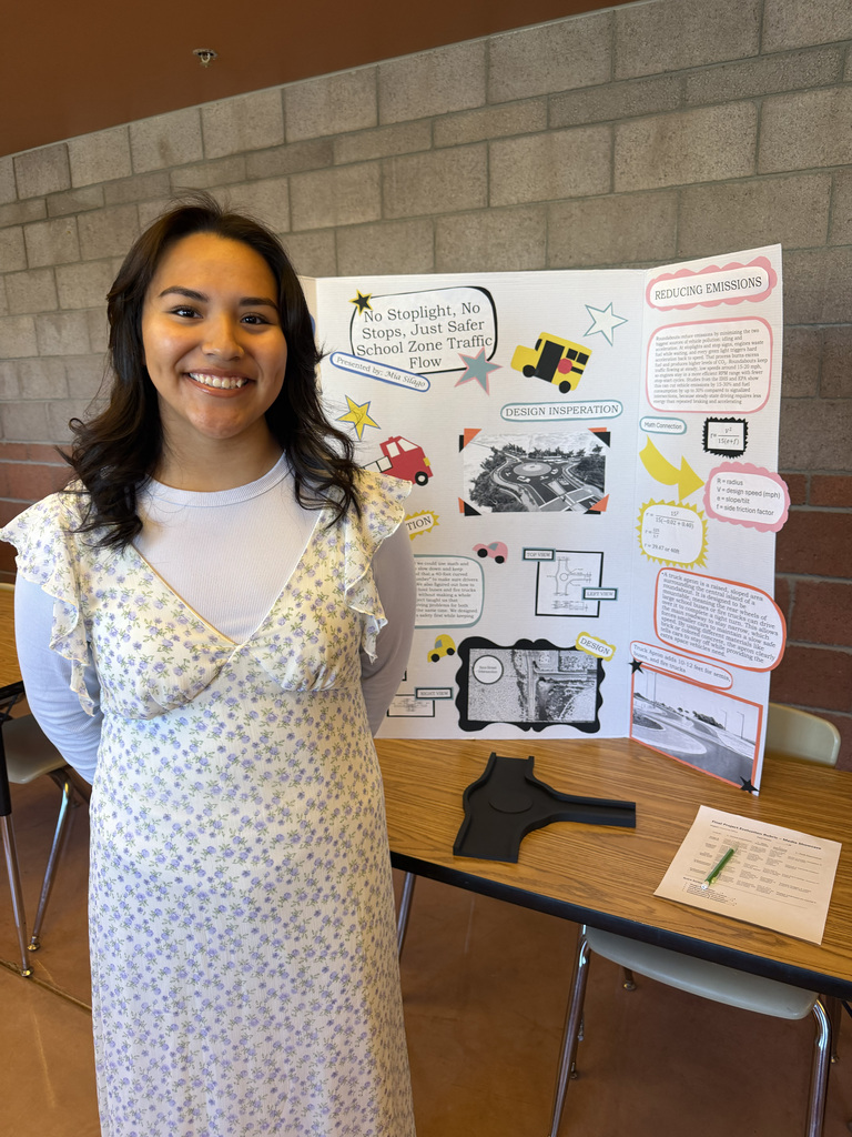A smiling woman in a floral dress stands beside a table displaying a poster and papers.