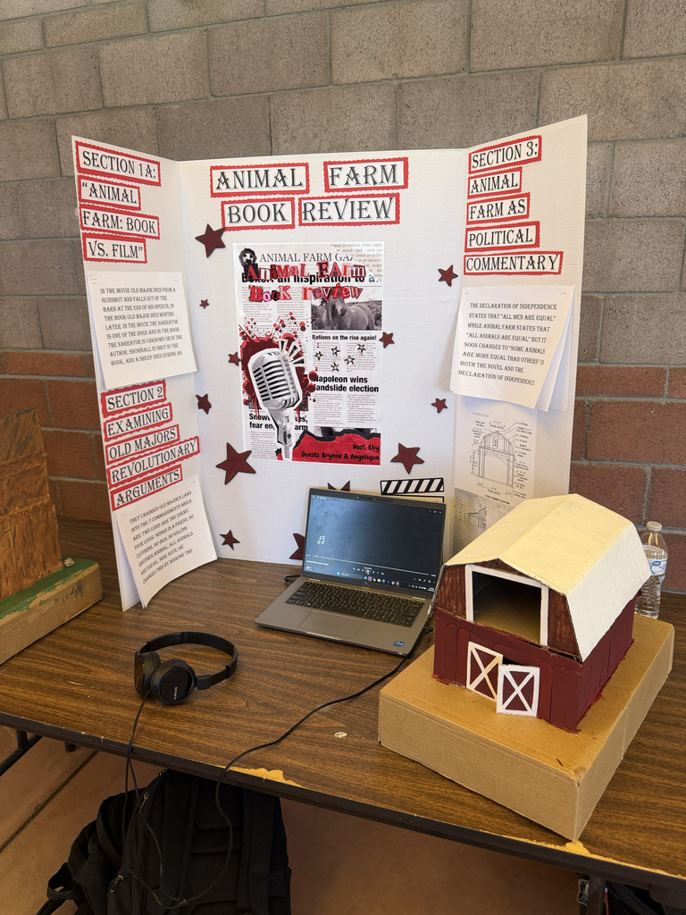 A desk displays a laptop, headphones, a bag, and a small barn model. A poster in the background has text about an Animal Farm book review.