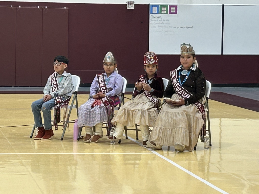 four students in traditional garb are seated in chairs in a gym