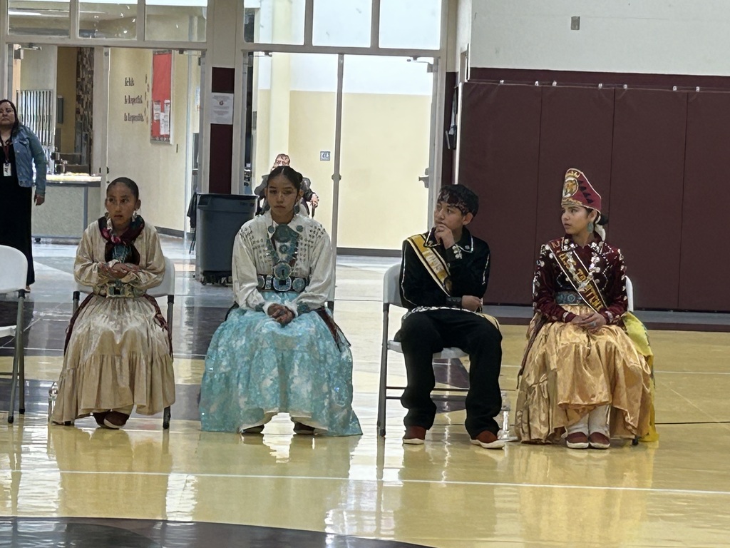 four students in traditional garb are seated in chairs in a gym