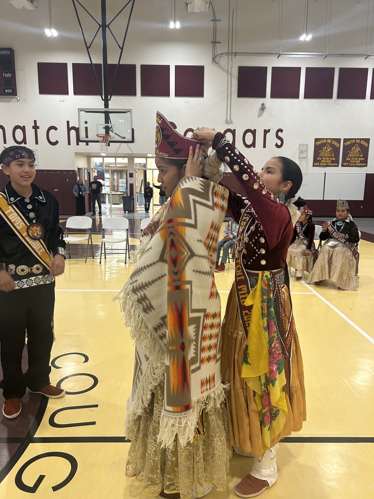 two girls are in traditional garb.  one is helping the  other put a crown on