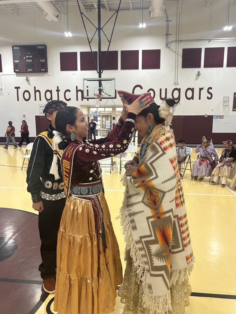 Two women in traditional garb are interacting on a basketball court, with a person in a black and gold dress and a man behind them.