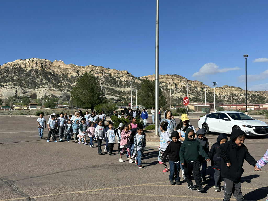 A group of children in various outfits walk in a line in a parking lot with mountains in the background.