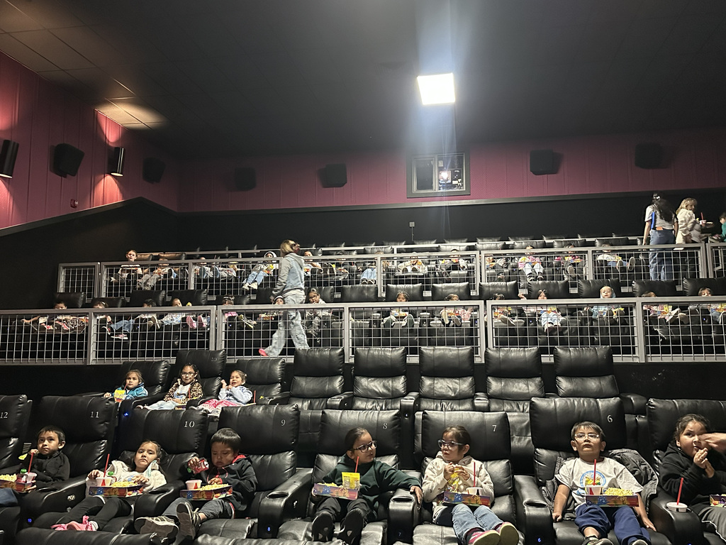 Interior of a movie theater with children seated in black chairs, a metal barrier, and pink walls.