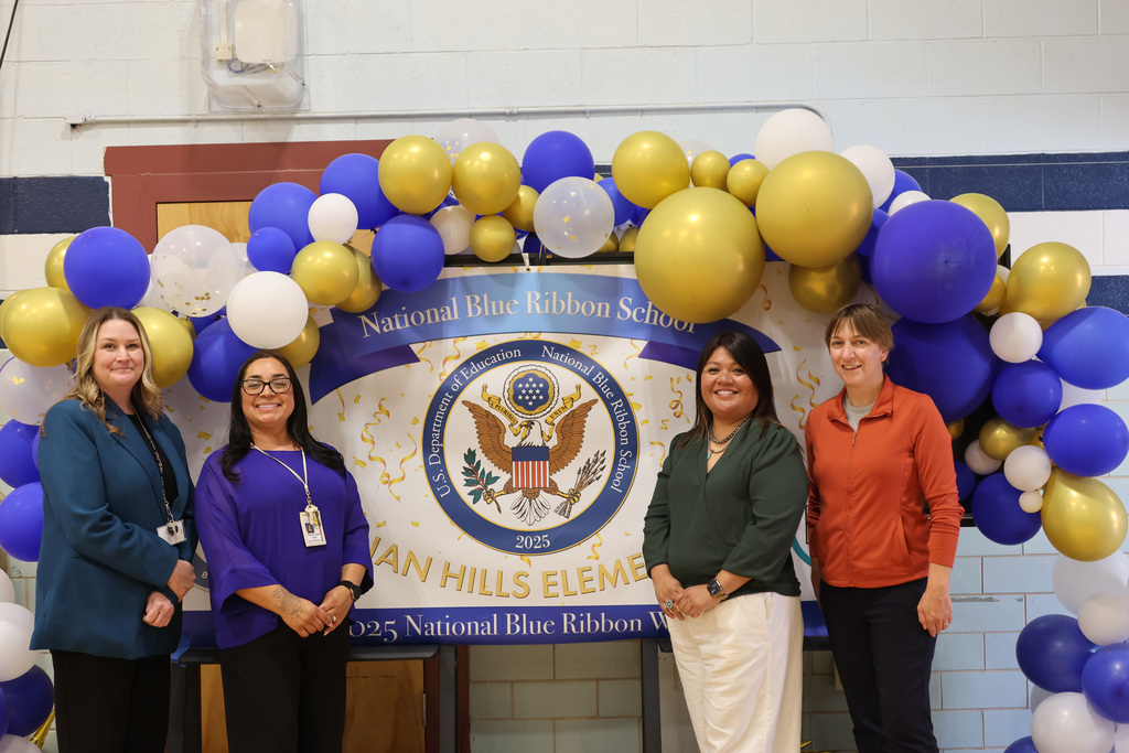 Five women in professional attire standing before a blue and gold balloon arch. Banner behind them reads "National Blue Ribbon School" with an emblem.