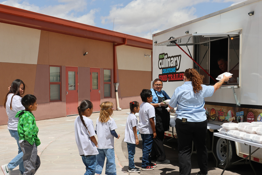 A food truck serves children, with two women in front. A woman hands out food. Children line up.