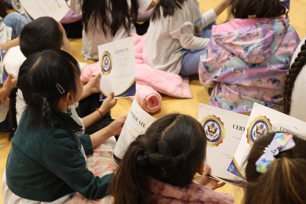 A group of children sit on the floor, holding certificates. Some have long hair and are wearing various clothes.