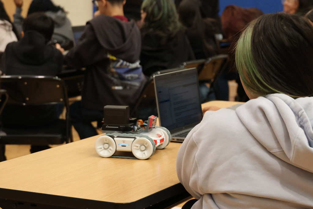 A student seated at a desk in a classroom, working on a laptop. A small robot with four wheels is on the desk.