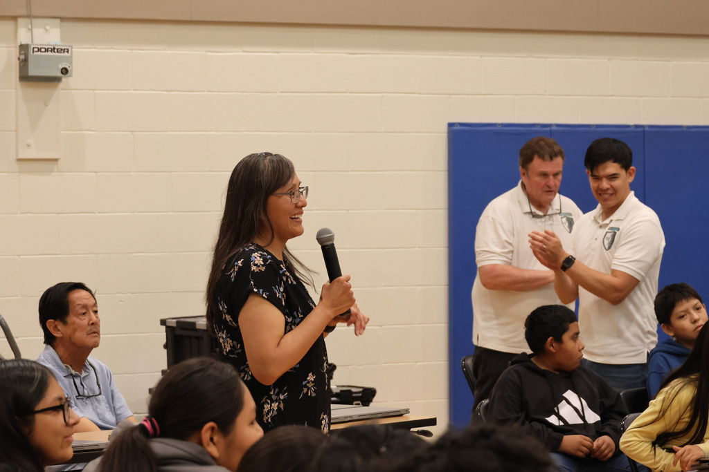 A woman speaks into a microphone in a classroom with several people seated in front of her.