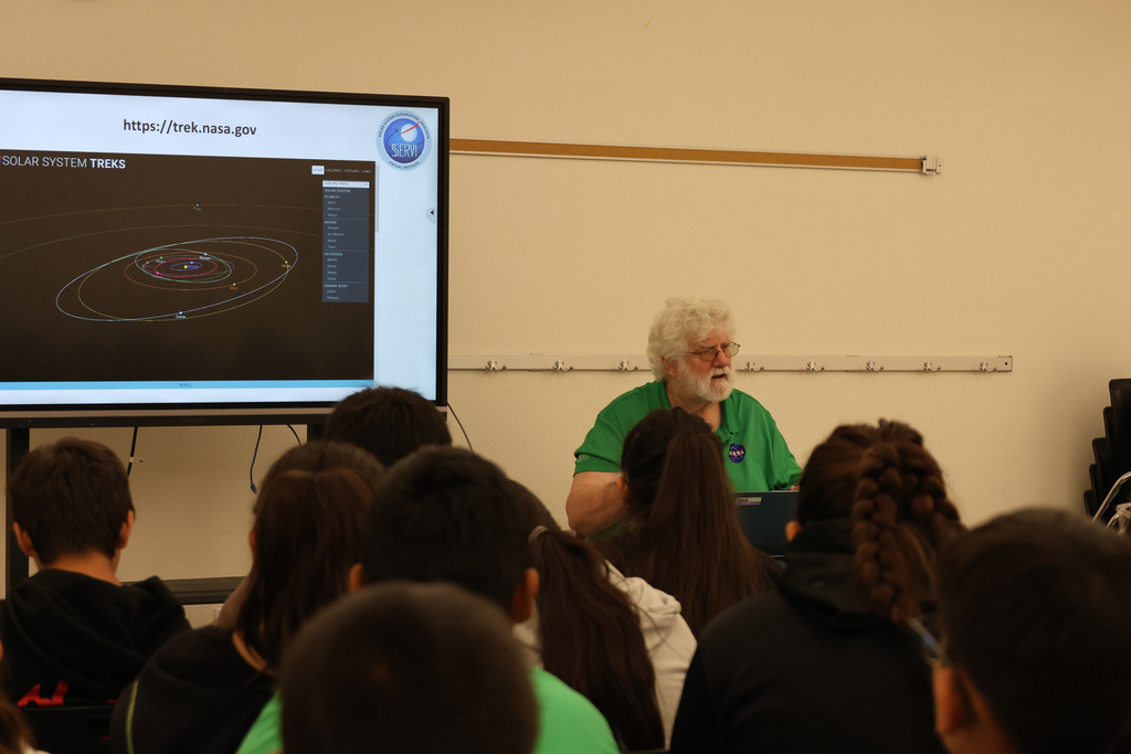 An elderly man in a green shirt speaks to a classroom of students, projected screen behind him.